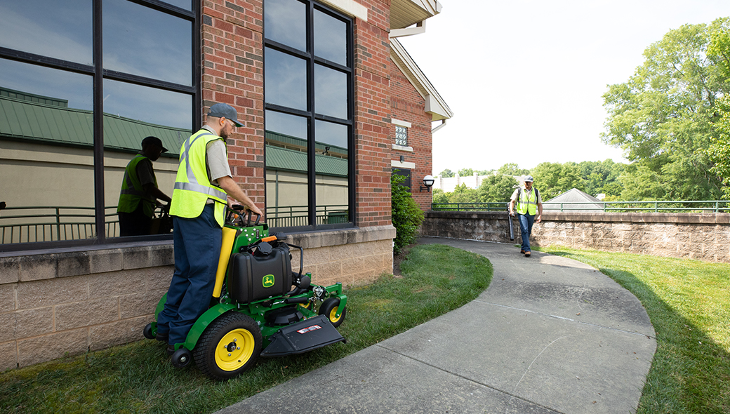 Man mowing in between a building and the sidewalk